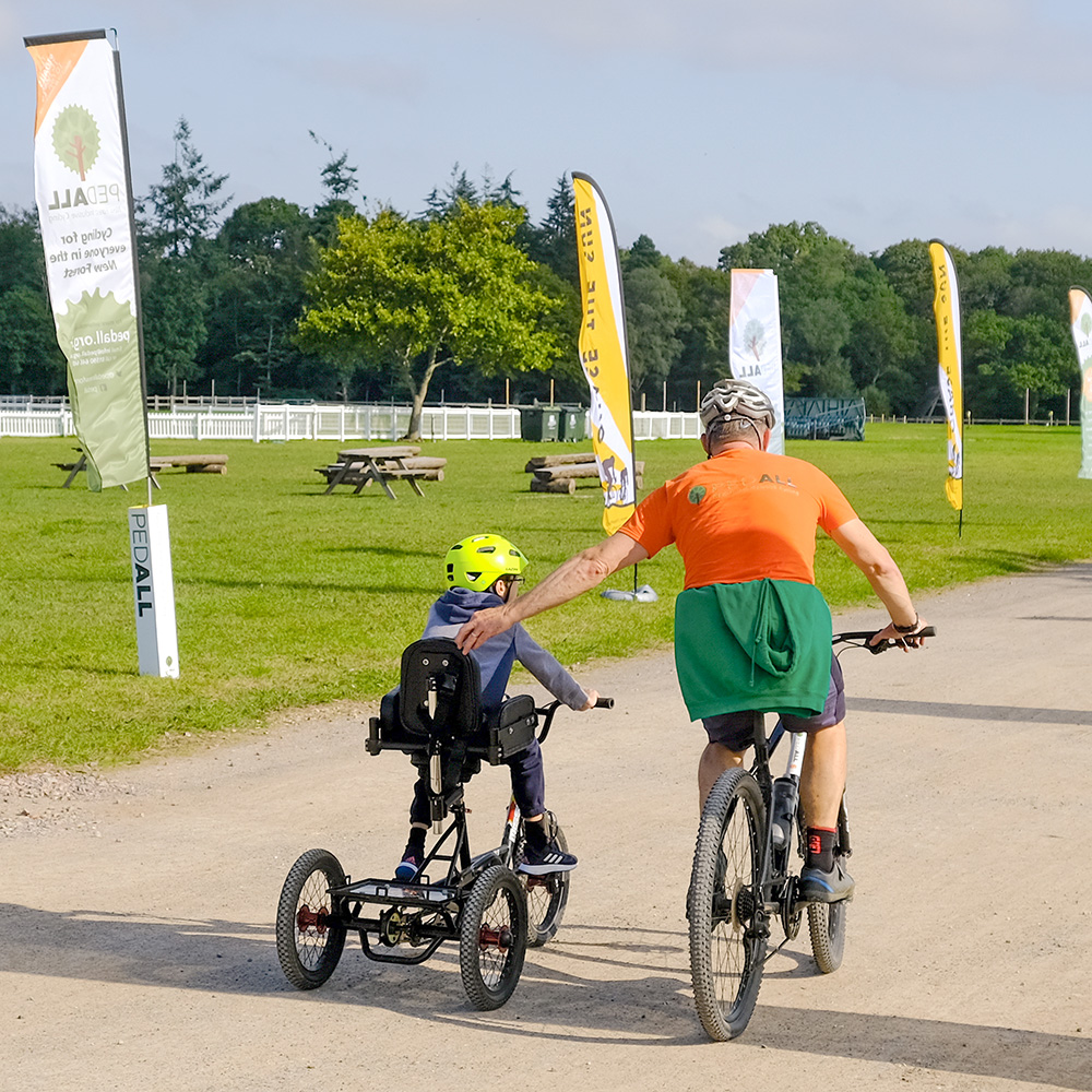 PEDALL volunteer on a bike helping a young child learn to cycle on a New Forest path