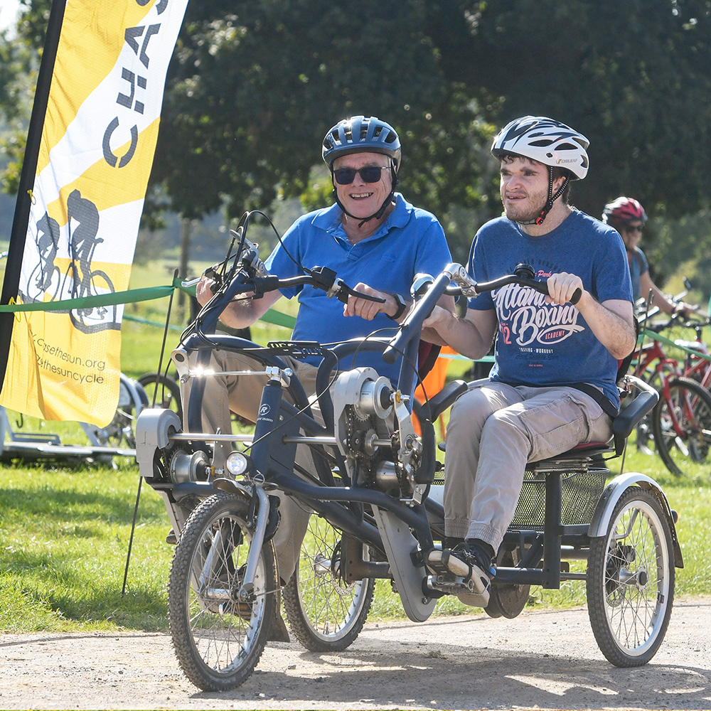 Two cyclists on a tandem bike smiling as they cycle during the Pass the PEDALL 2024 event, in front of a banner